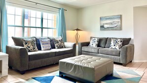 A living room with two grey sofas, a white ottoman, a lamp, and a painting of a boat on the wall. - 🏖️BEACHSIDE🏖️ 2/2 Condo In The Heartbeat of SPI!! (South Padre Island)