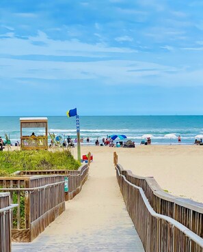 Une promenade en bois mène à une plage où se trouve un poste de surveillance des sauveteurs, des parasols et des personnes qui profitent du bord de mer.