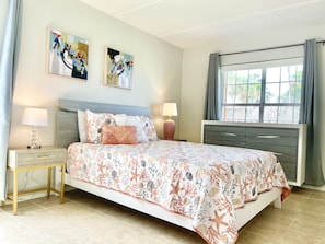 Un dormitorio con cama, mesitas de noche, cómoda y dos cuadros en la pared, además de una ventana con cortinas.
