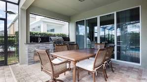 An outdoor patio with a wooden table and wicker chairs, a grill, and a glass-enclosed area with a view of a swimming pool. - Fantabulous Disney Vacation House - Private pool, Movie Theater, Game Room (Kissimmee)