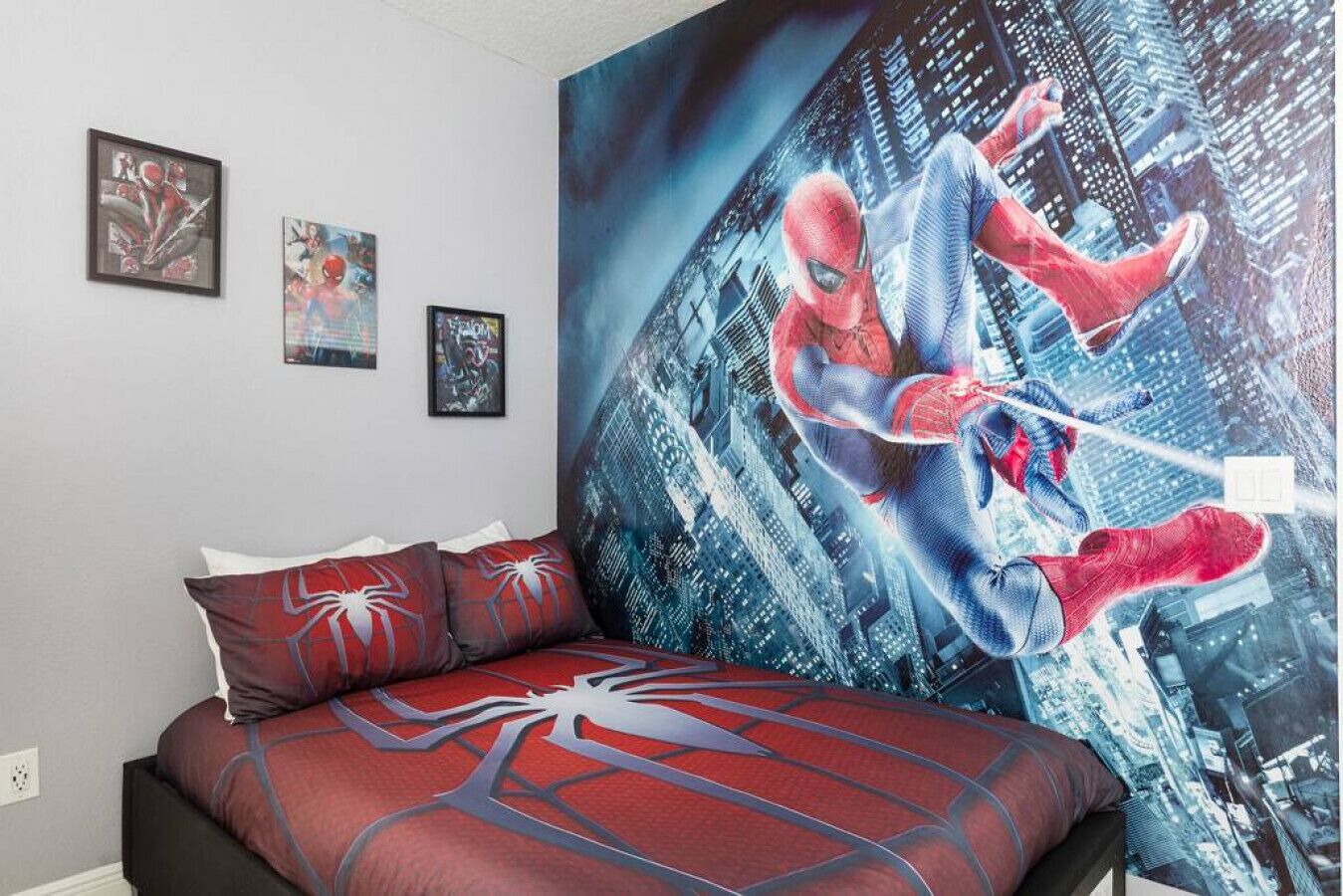 A bedroom with a large Spider-Man mural on the wall, a bed with Spider-Man themed bedding, and two framed posters on the wall.