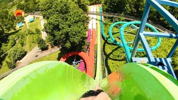 A person sliding down a bright green water slide with a view of a roller coaster and trees in the background.