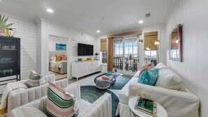 A modern living room with a sofa, armchairs, a coffee table, and a TV. There is a dining area with a table and chairs visible through a glass door. - Newly Renovated Professionally Decorated Gulf Views! Pool & Beach Access. (Seacrest Beach)