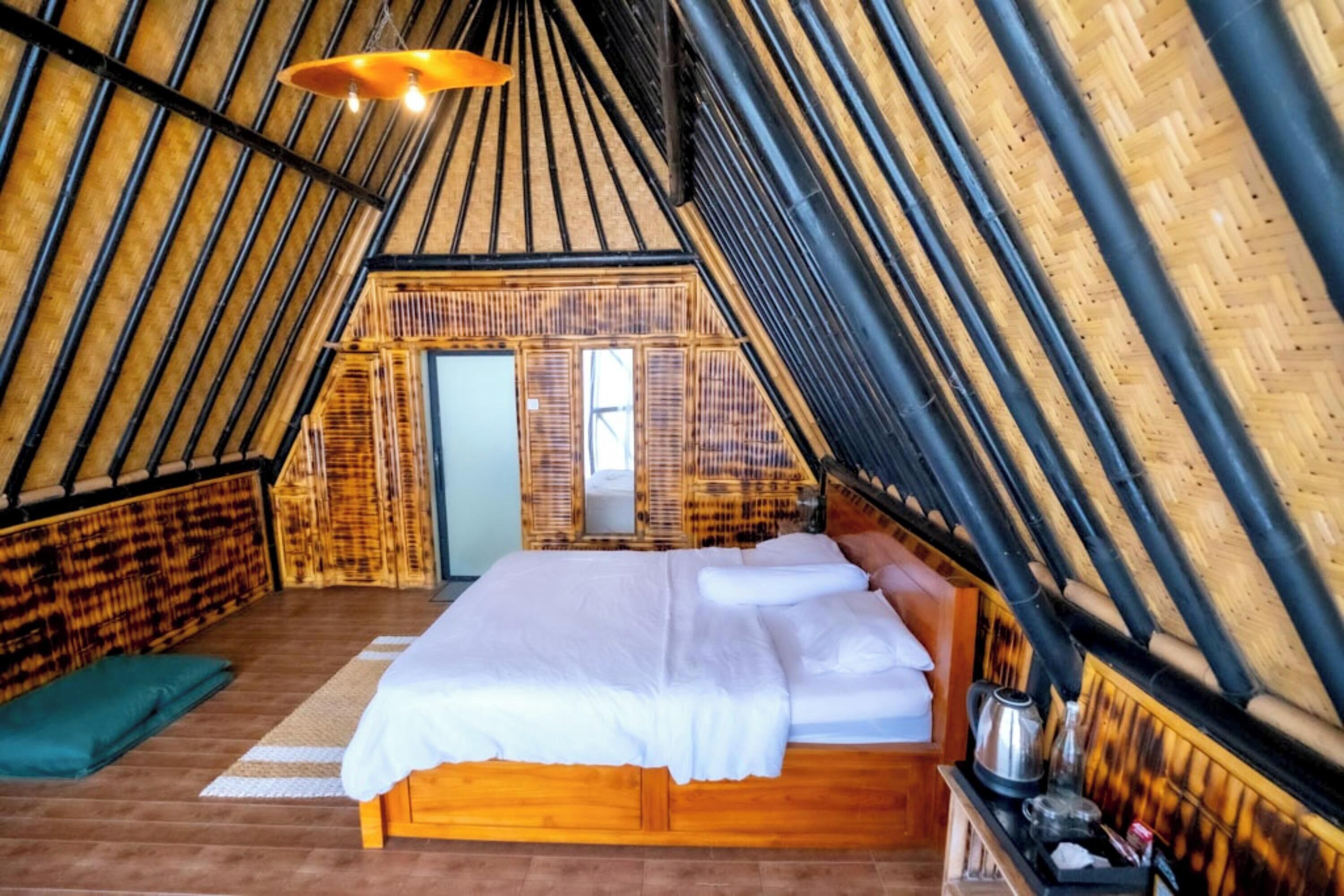 A cozy, triangular-roofed room with a wooden bed, a small table with a kettle, and a wicker chair.