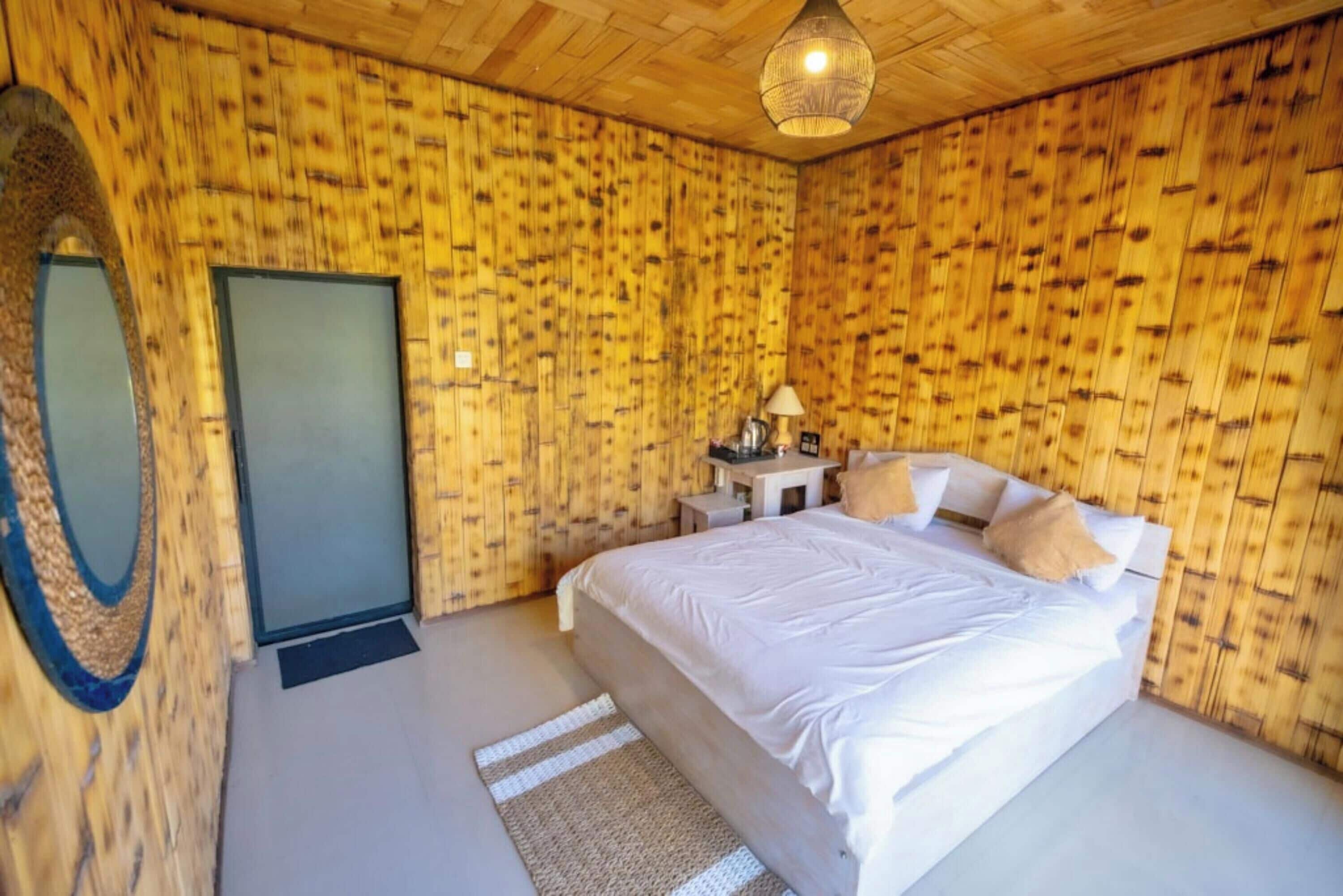 A bedroom with a wooden wall panel design, a bed with white linens, a bedside table with a lamp, and a large mirror on the left wall.