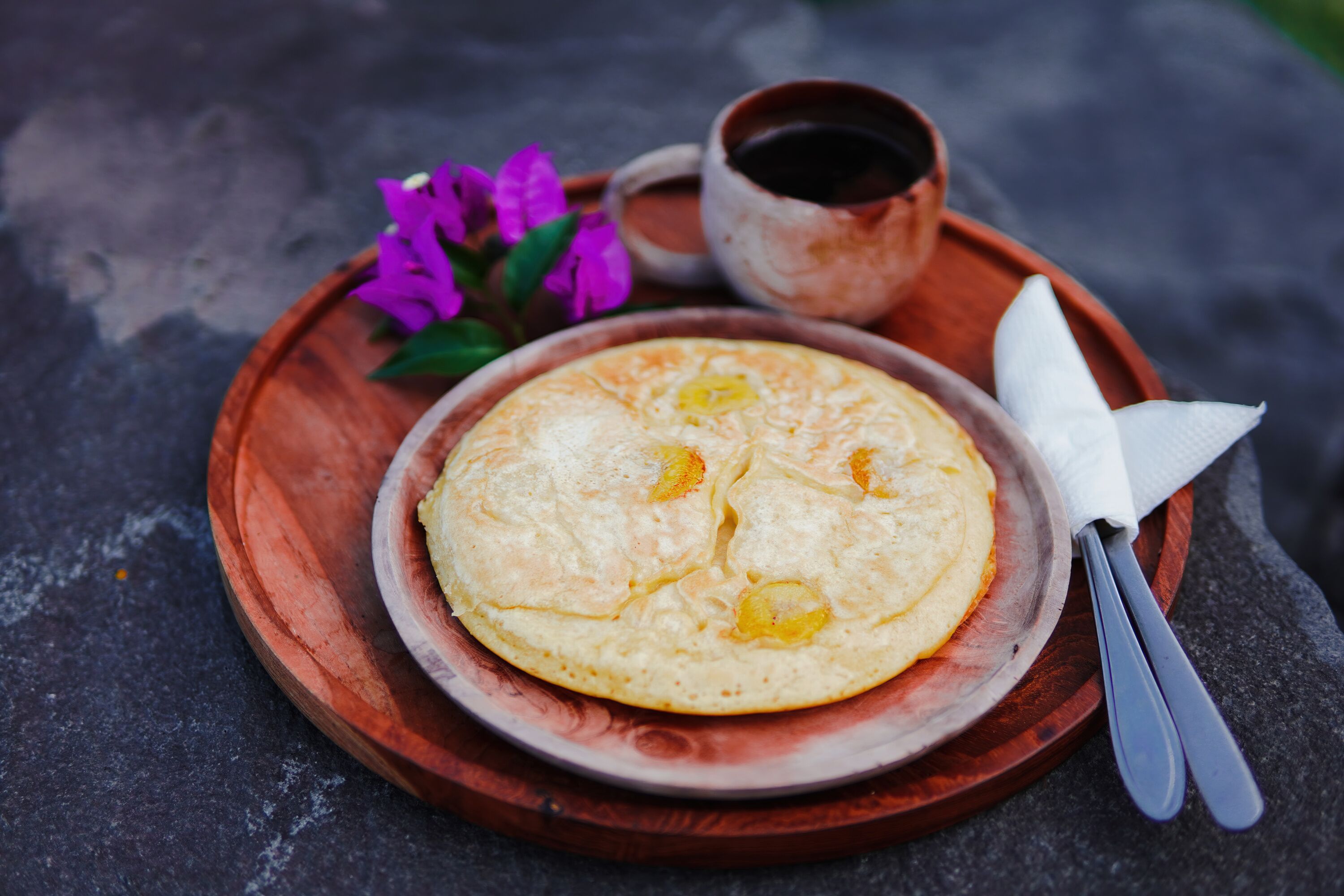 A wooden plate with a breakfast dish, a cup of coffee, and a napkin on a stone surface.