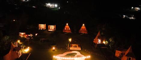 A nighttime view of a resort with multiple bungalow-style accommodations, a central illuminated pathway, and surrounding greenery.