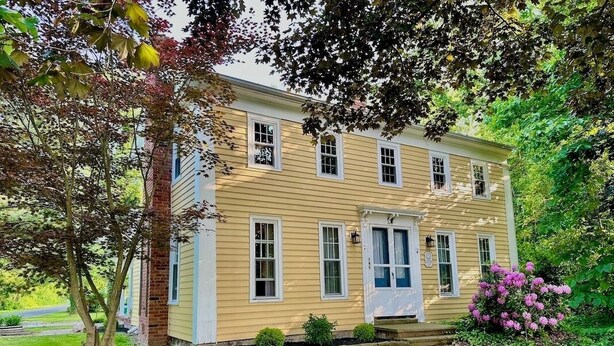 A two-story yellow house with a front porch, a brick chimney, and a small garden with a tree and flowering bushes.