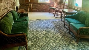 A cozy room with wooden ceiling beams, a brick wall, and a patterned floor. There are two green sofas and a rocking chair.