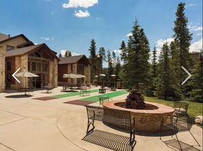 Relax by the stone fireplace in a serene forest setting, surrounded by comfortable seating and shaded by umbrellas.