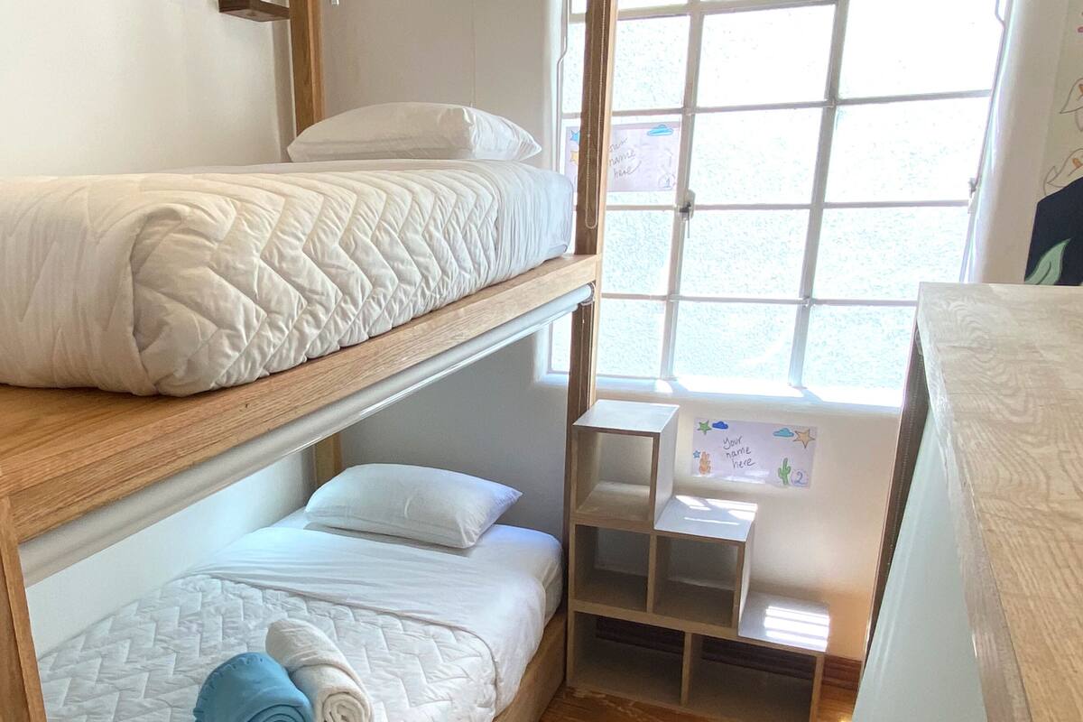 A bunk bed room with a wooden frame, a small desk with a name tag, and a window with a view.