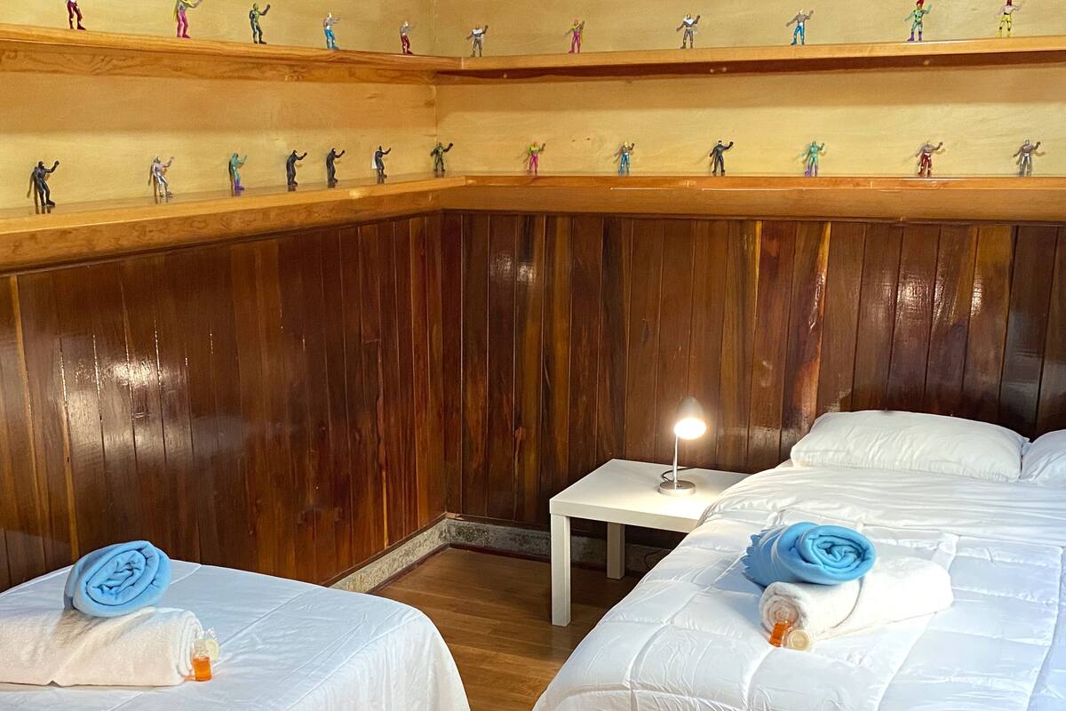 A hotel room with wooden paneling, two beds with white linens, a small bedside table with a lamp, and a shelf with figurines.