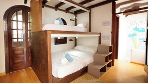 Una cama litera con estructura de madera, un colchón blanco y una almohada azul. Se observa una puerta de madera con paneles de vidrio y una ventana pequeña. Una pared blanca muestra un interruptor de luz y una estantería fijada a la pared.