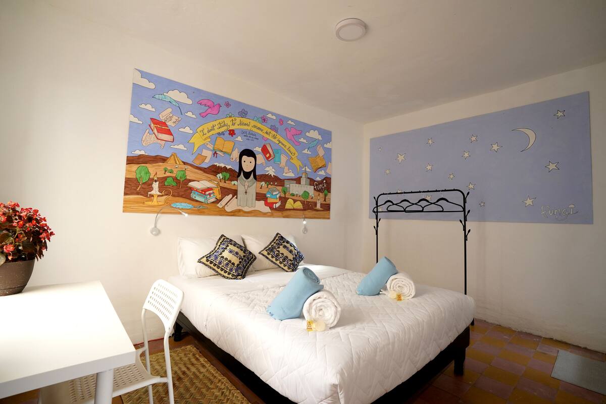 A bedroom with a bed, pillows, a bedside table, a chair, a wall mural, and a starry night wall decal.