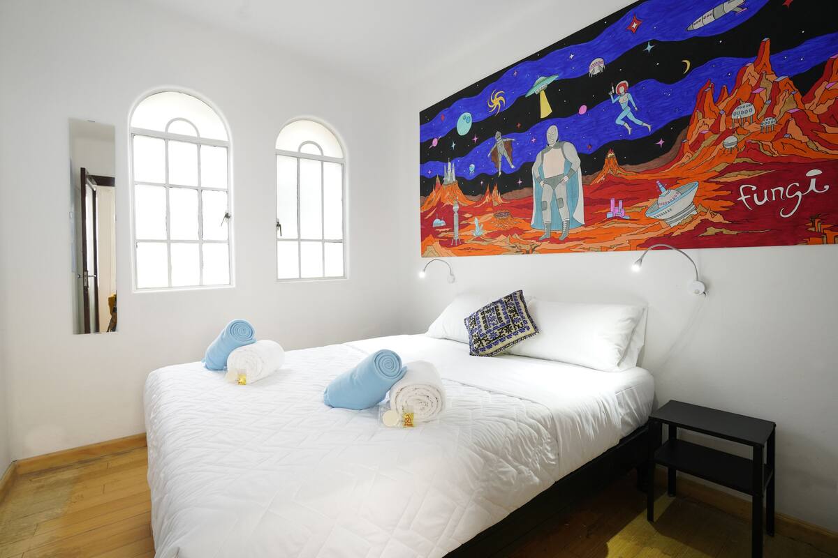 A bedroom with a bed, two windows, a painting, and a nightstand.