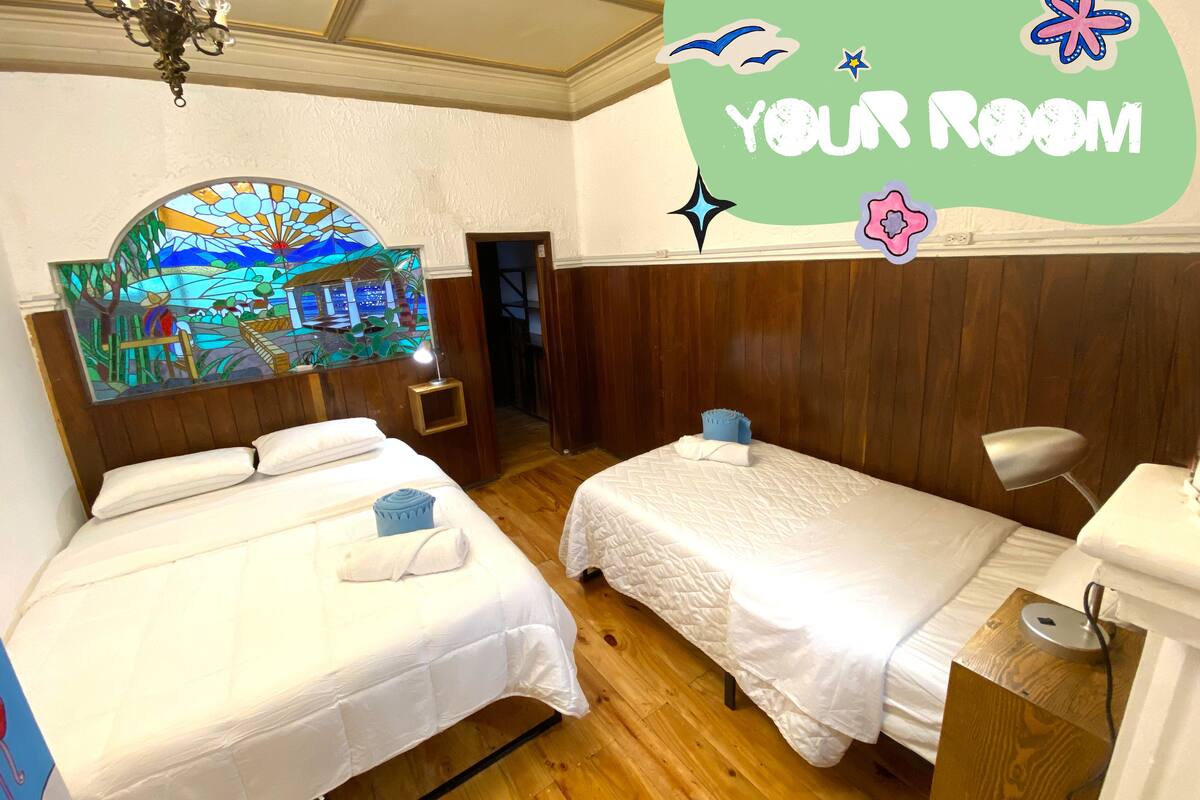 A hotel room with two beds, a wooden headboard, a stained glass window, and a wall decoration reading "YOUR ROOM".
