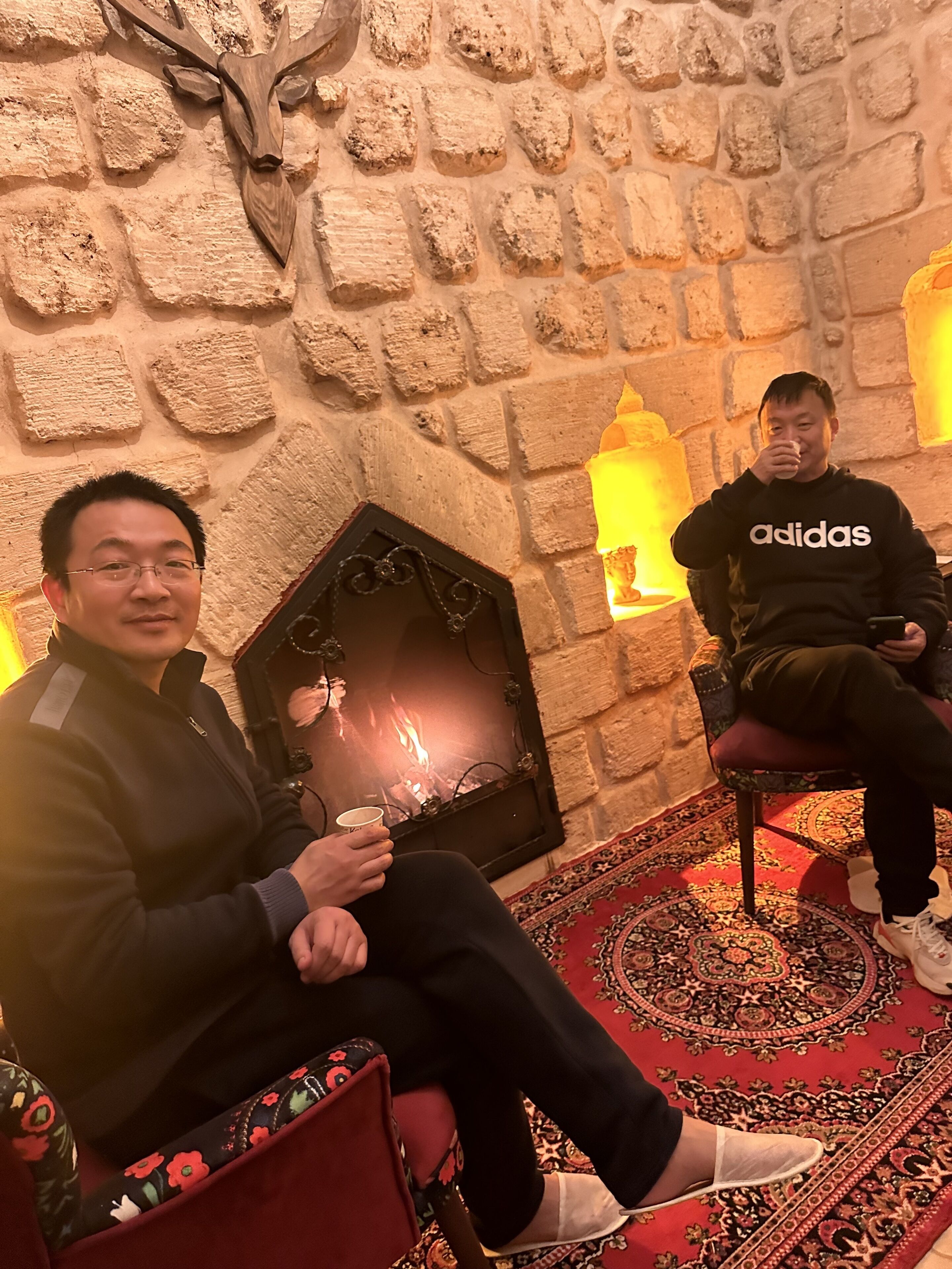 Two people seated in a room with a stone wall, a fireplace, and patterned carpet.