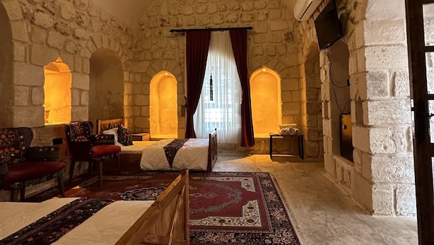 A spacious room with a high arched stone ceiling, a wooden bench, a bed with a red and white patterned rug, and a view through a window with red curtains. - Şikeft Butik Otel (Midyat)