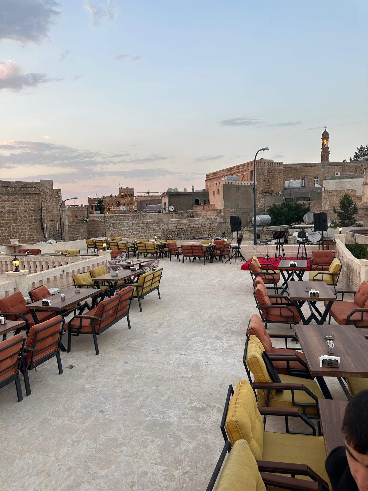 An outdoor seating area with tables and chairs, stone walls, and a view of historic buildings in the background.