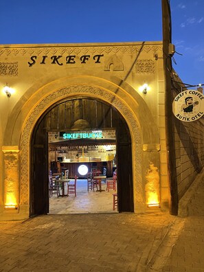 L’entrée d’un immeuble, avec une porte en arc de cercle, une enseigne indiquant « SIKEFT COFFEE BUTIK OTEL » et un espace bar à l’intérieur, avec des tables et des chaises.
