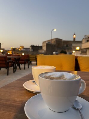 A cup of coffee on a table with a spoon, in an outdoor seating area with chairs and tables, and a cityscape in the background. - Şikeft Butik Otel (Midyat)