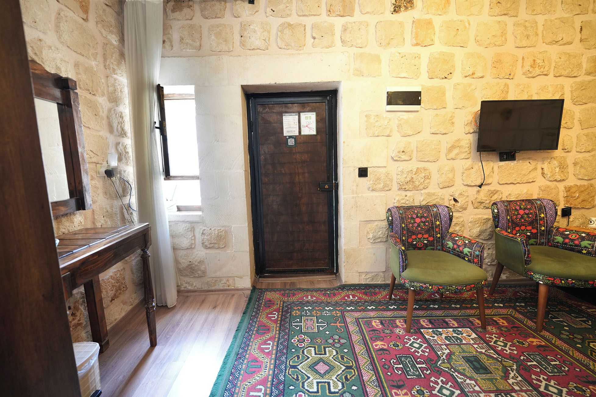 A room with a stone wall, a wooden door, two patterned chairs, a wooden bench, and a television mounted on the wall.