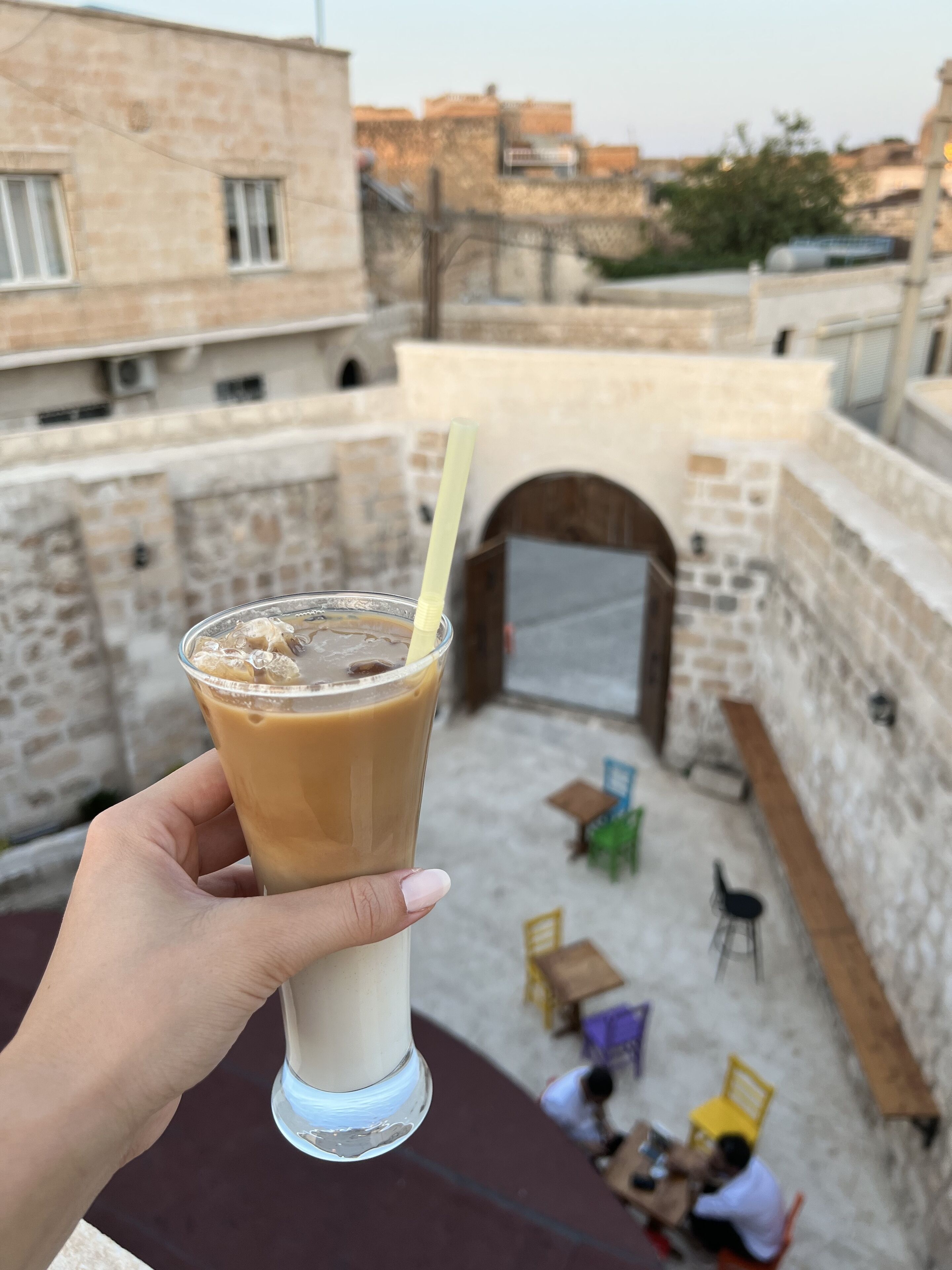 Una mano sostiene un vaso de café helado con pajita, con vista a un patio con muros de piedra, un banco de madera y algunas personas sentadas en mesas.