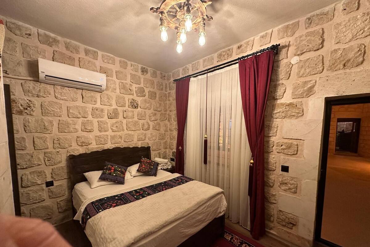 A bedroom with a stone wall, a bed with patterned pillows, a chandelier, and a red curtain.
