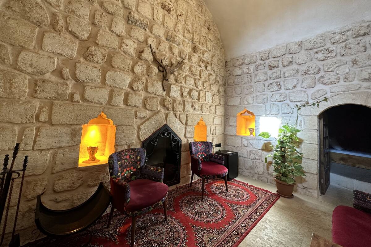A stone-walled room with a fireplace, two chairs, a patterned rug, and a potted plant.