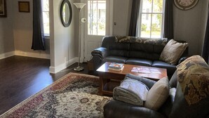 A living room with a dark leather sofa, a patterned rug, a coffee table, a floor lamp, and a large window with curtains. - Beautiful 4-Bedroom Home • Walk to Beach, Riverwalk & Downtown (Manistee)