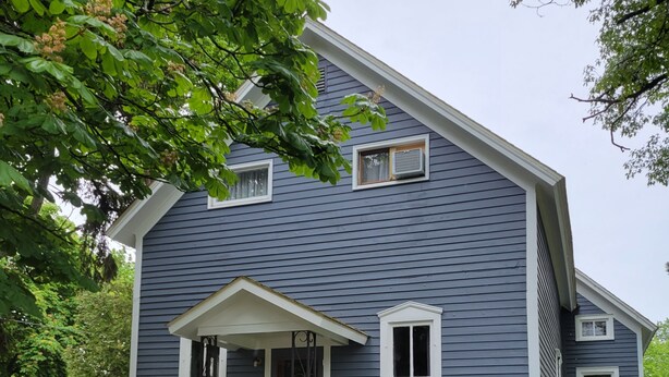 A two-story house with a front porch, a small front yard with flowers, and a driveway. - Beautiful 4-Bedroom Home • Walk to Beach, Riverwalk & Downtown (Manistee)