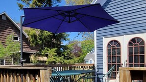 A wooden deck with outdoor furniture, a blue umbrella, and a house with a blue exterior and arched windows. - Beautiful 4-Bedroom Home • Walk to Beach, Riverwalk & Downtown (Manistee)