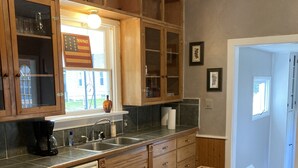 A kitchen with wooden cabinets, a dishwasher, a window with a view, and a table with a placemat. - Beautiful 4-Bedroom Home • Walk to Beach, Riverwalk & Downtown (Manistee)