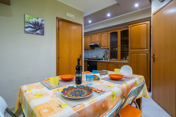 Un comedor con una mesa preparada para cuatro personas, sillas anaranjadas, mantel estampado y una botella de vino.