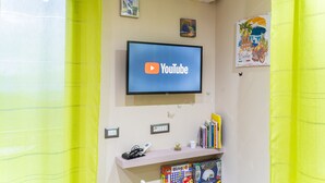 A room with a flat-screen TV mounted on the wall displaying the YouTube logo, a small shelf with books and a phone, and a window with yellow curtains. - Angel's Holiday Apartment - Panoramic Relax (San Mauro Castelverde)