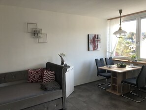 Un salon moderne avec un canapé gris, des coussins à motifs, une table à manger avec des chaises et une vue sur les maisons par la fenêtre.