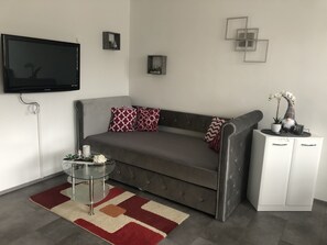 Un salon moderne avec un canapé gris, des coussins à motifs, une table basse en verre, un meuble blanc et un téléviseur à écran plat fixé au mur.
