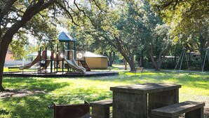Ein Spielplatz mit Rutschen und Schaukeln, umgeben von Bäumen und einem Picknicktisch.