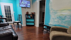 Ein Wohnzimmer mit Holzboden, einer schwarzen Ledersofa, einem beigefarbenen Sessel, einem Glastisch und einem an der Wand montierten Fernseher.
