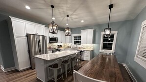 A modern kitchen with white cabinets, stainless steel appliances, a wooden dining table, and pendant lighting.