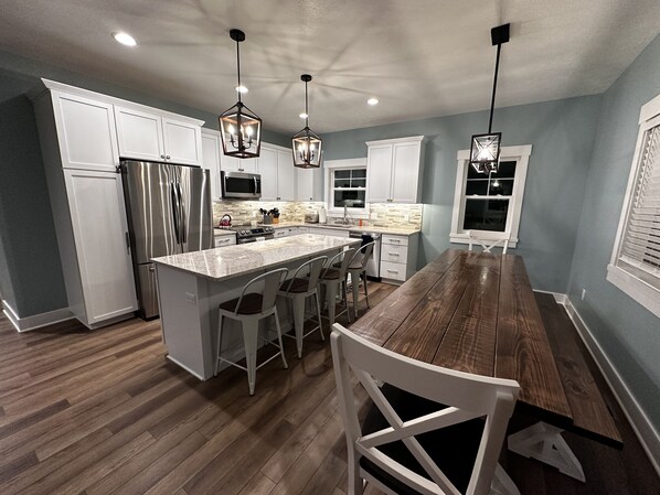A modern kitchen with white cabinets, stainless steel appliances, a wooden dining table, and pendant lighting. - Luxury house, covered patio, private fire pit patio. Memorial Day Weekend Open (Mears)