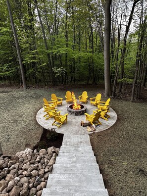 A circular outdoor seating area with yellow chairs around a central fire pit, surrounded by a forest. - Luxury house, covered patio, private fire pit patio. Memorial Day Weekend Open (Mears)