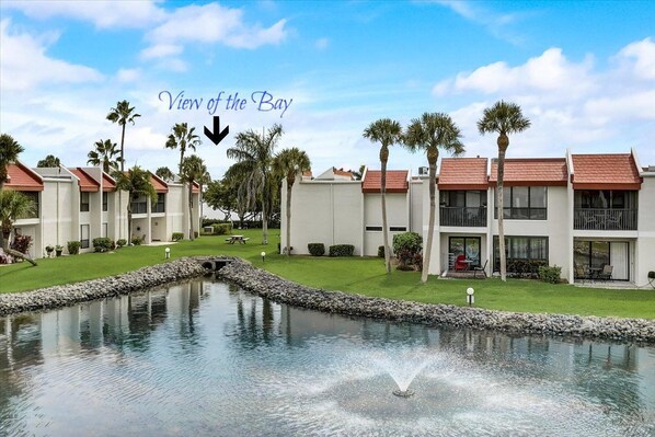 A resort with a view of the bay, featuring a pool with a fountain, palm trees, and modern buildings with red roofs.