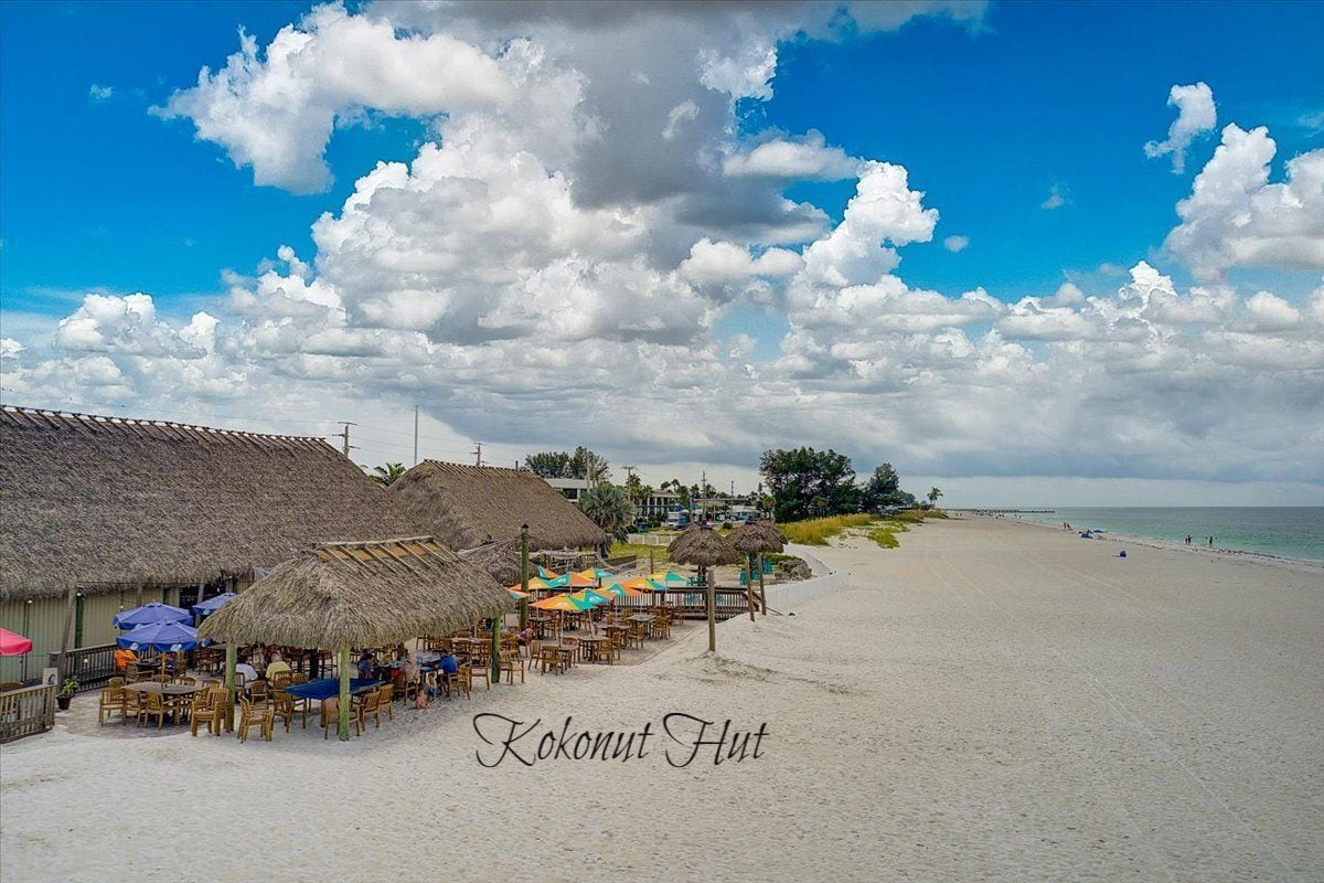 A beachside restaurant with thatched roofs and outdoor seating, located on a sandy beach with a view of the ocean.