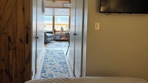 Ein Schlafzimmer mit Bett, einem an der Wand befestigten Fernseher und Blick auf einen Balkon mit Geländer und einem Schild mit der Aufschrift 'Bitte hier bleiben'.