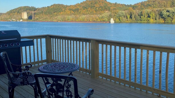 Eine Holzterrasse mit Tisch und Stühlen mit Blick auf einen See, im Hintergrund ein Boot und eine Brücke.