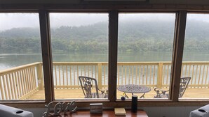 Ein Zimmer mit großem Fenster mit Blick auf einen See und einen Wald. Es steht ein Holztisch mit einem Topfpflanzen, ein Stuhl und ein Schild mit der Aufschrift 'Love'.