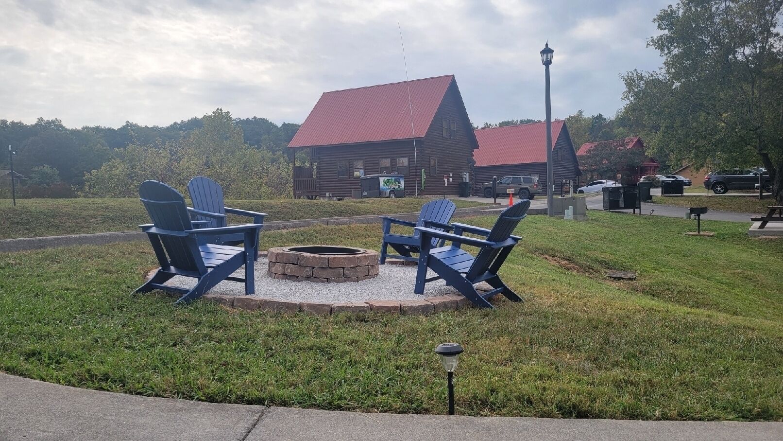 A wooden cabin with a red roof, a fire pit with chairs, and a lamp post.