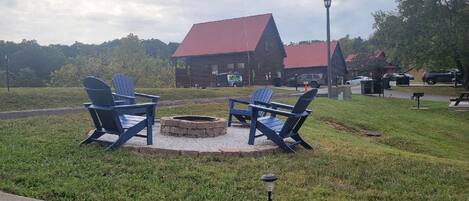 A wooden cabin with a red roof, a fire pit with chairs, and a lamp post.
