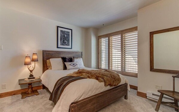 Quarto com cama de madeira, mesa de cabeceira com luminárias, quadro na parede e janela com persianas.